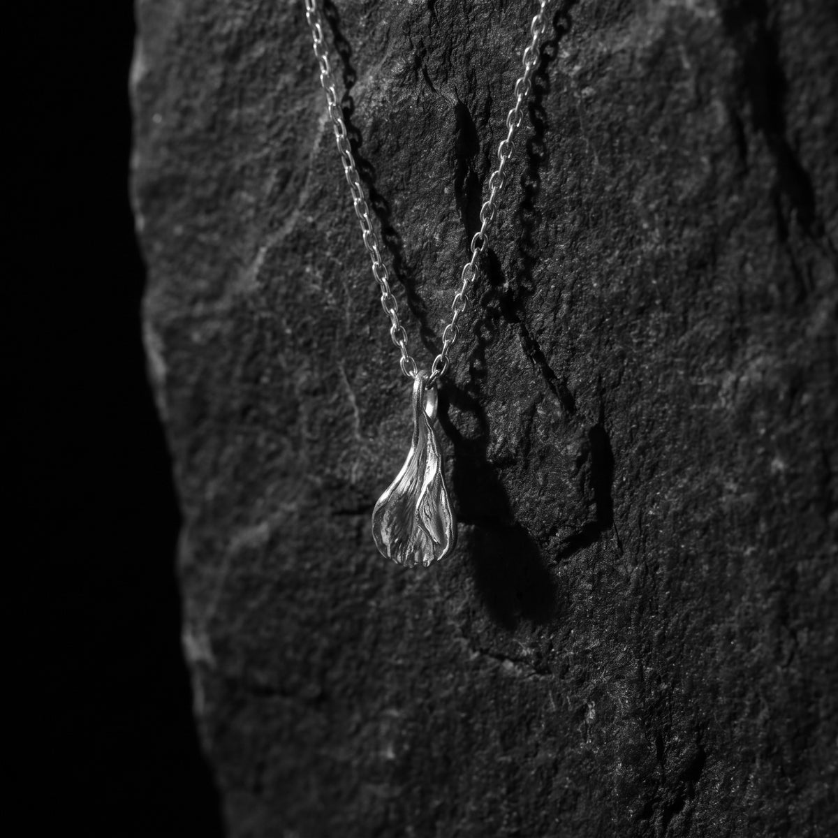 Vertical view of the textured silver petal pendant hanging against dark textured rock.