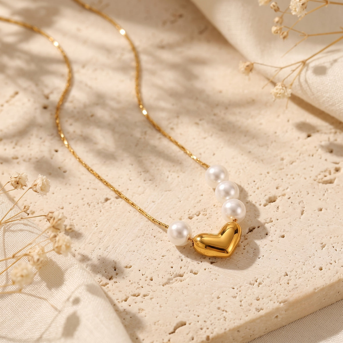 Gold-tone heart and asymmetrical pearl necklace lying flat on textured stone beside dried flowers.