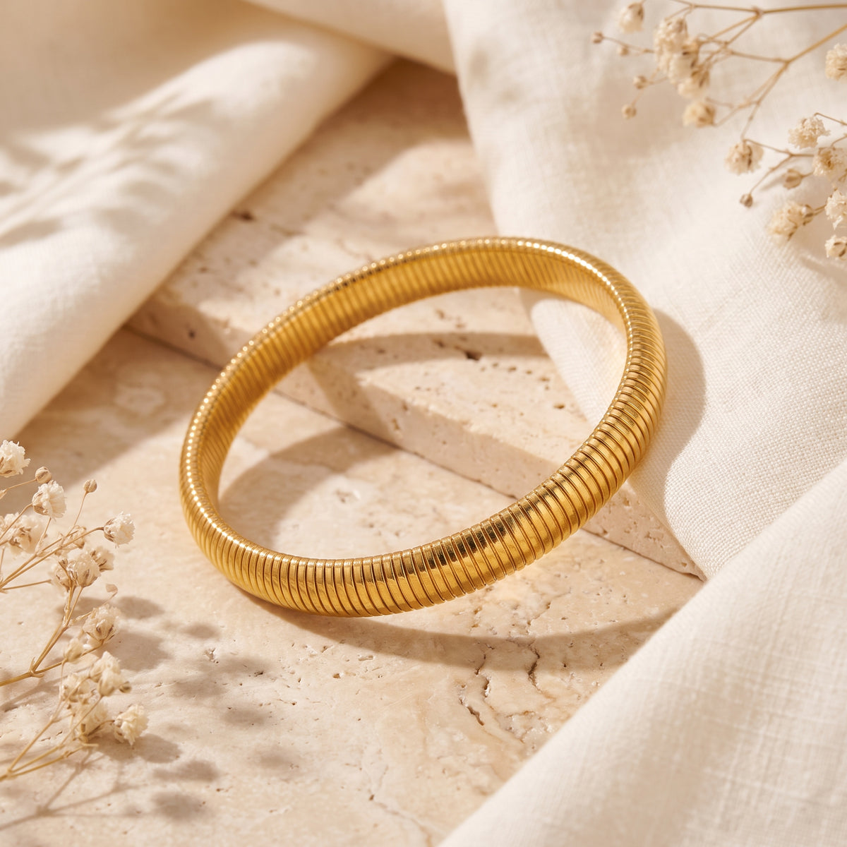 Angled gold-tone ribbed bracelet resting across a travertine block beside cream fabric and dried flowers.