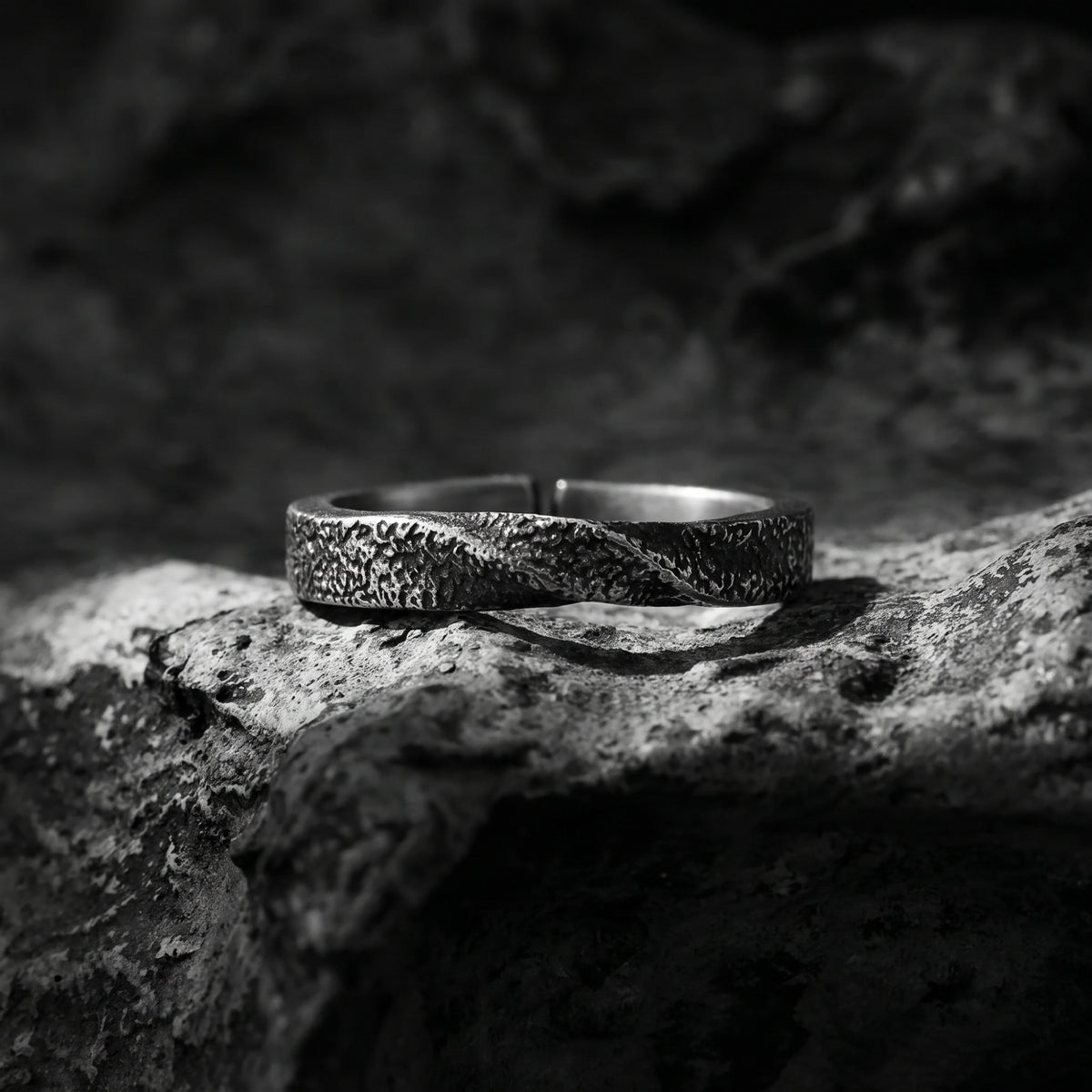 Dark textured open-band ring with a diagonal twist resting on rough stone.