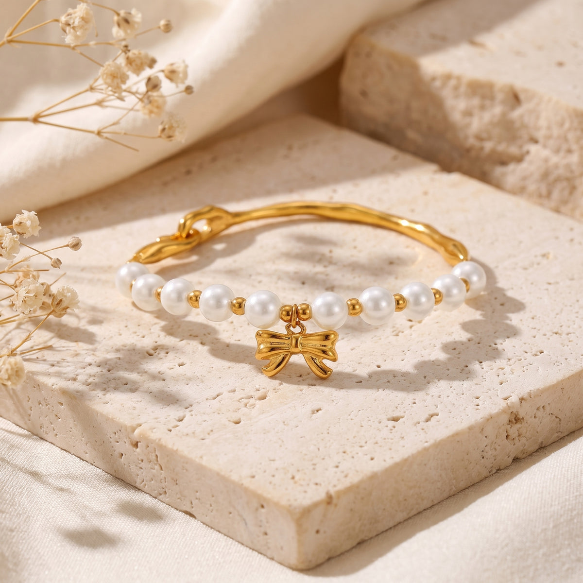 Angled view of the pearl bracelet on a porous beige stone slab, with the gold-tone bow charm centered.