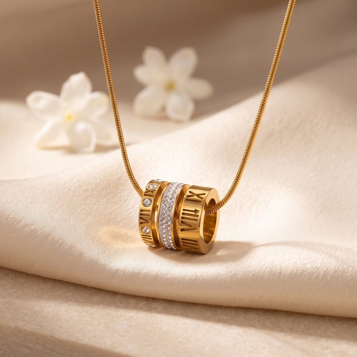 Gold-toned Roman numeral barrel pendant necklace resting on draped cream fabric with white flowers.