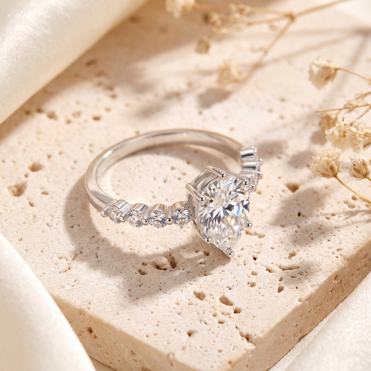 Side angle view resting on a beige slab, showing the ring's pear-cut center stone and prong-set side stones.