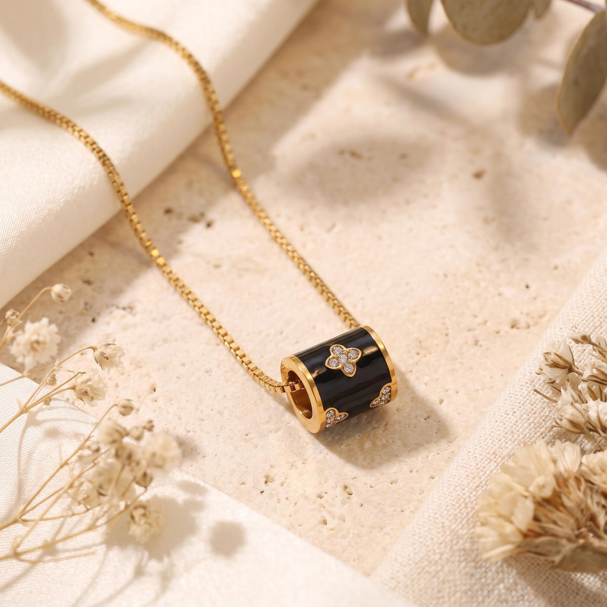Flat-lay gold-tone necklace with a black enamel barrel pendant on a light stone surface among dried flowers.