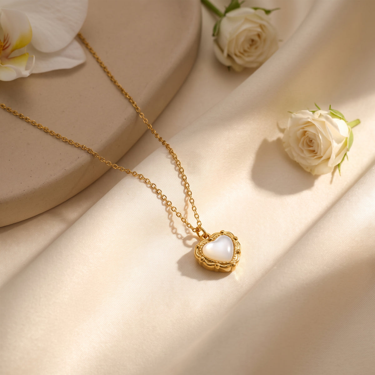 Gold-tone heart pendant necklace with a white mother-of-pearl simulant inlay laid across satin beside white roses.