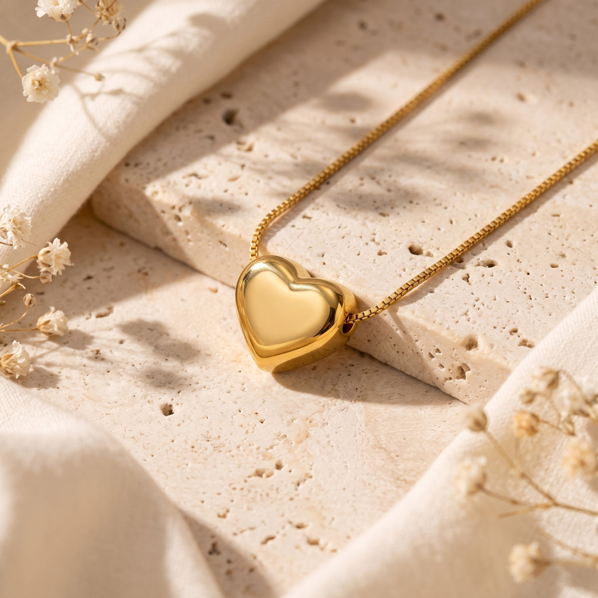 Gold-tone puffy heart pendant necklace laid diagonally on a porous stone slab with dried flowers.