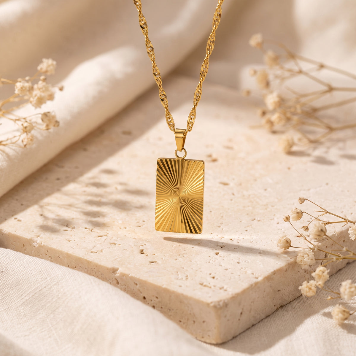 Gold-tone sunburst plaque pendant hanging above a beige textured slab, framed by dried flowers.