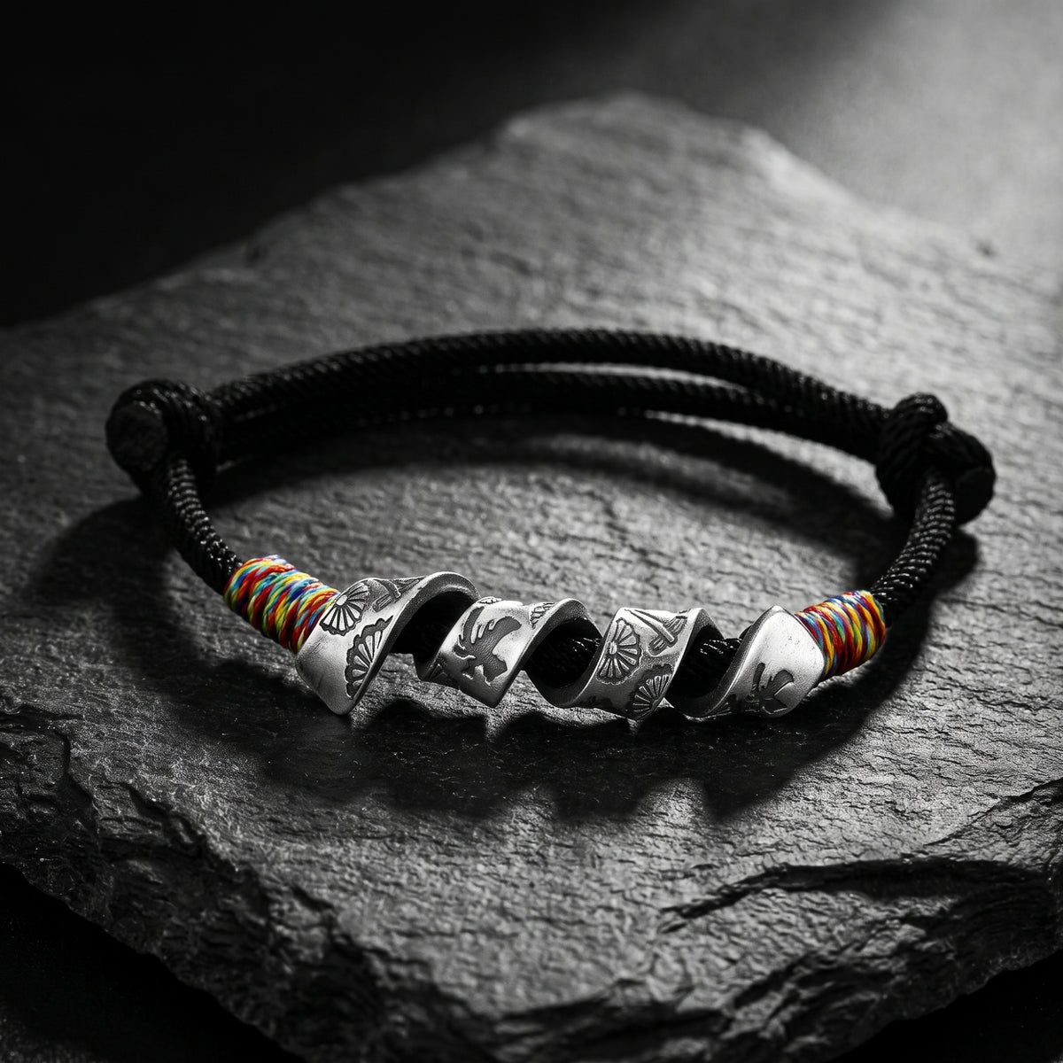 Overhead view of a black cord bracelet with multiple engraved silver sections and colorful thread accents on slate.