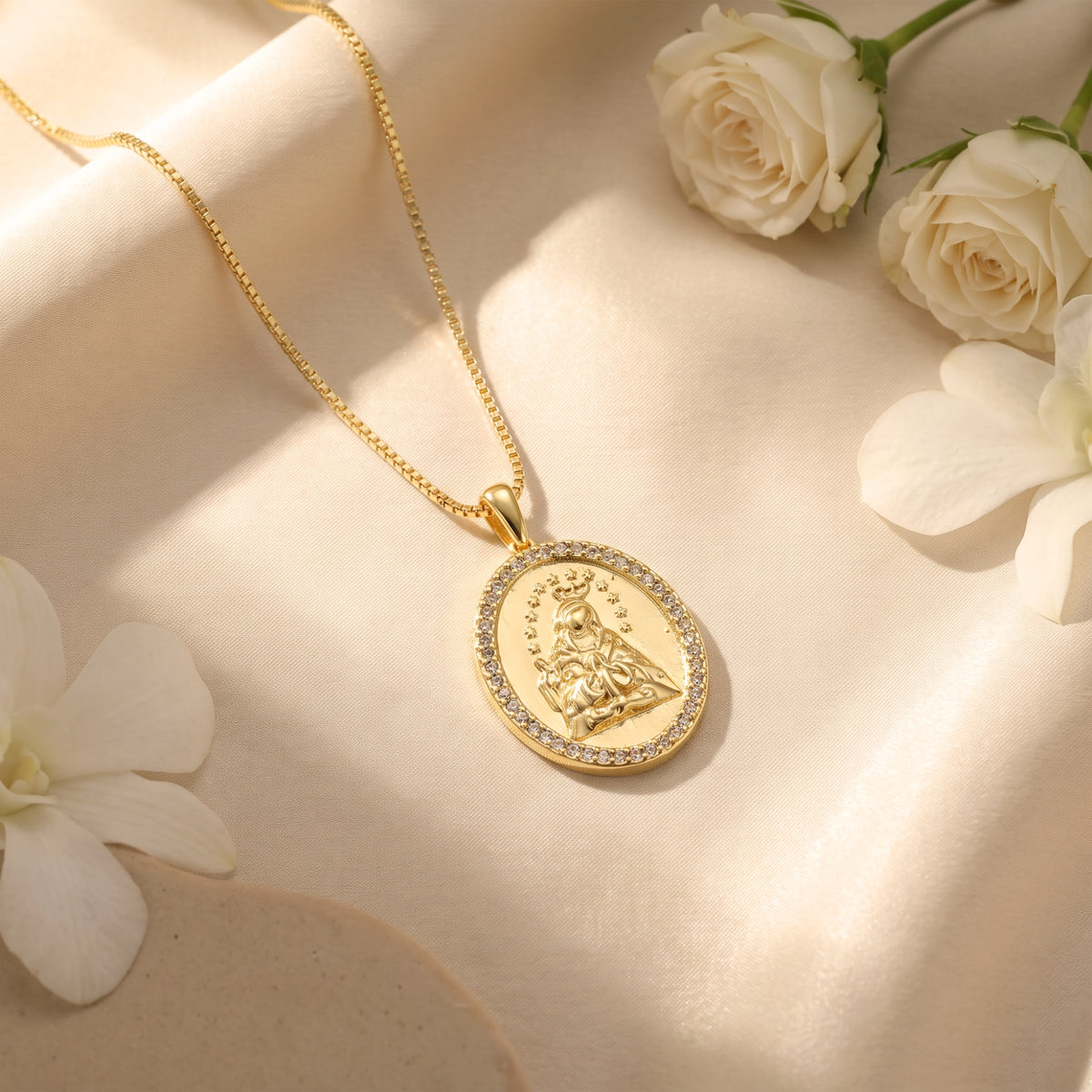 Gold-tone Marian medallion necklace with a rhinestone border resting on beige satin beside white roses.
