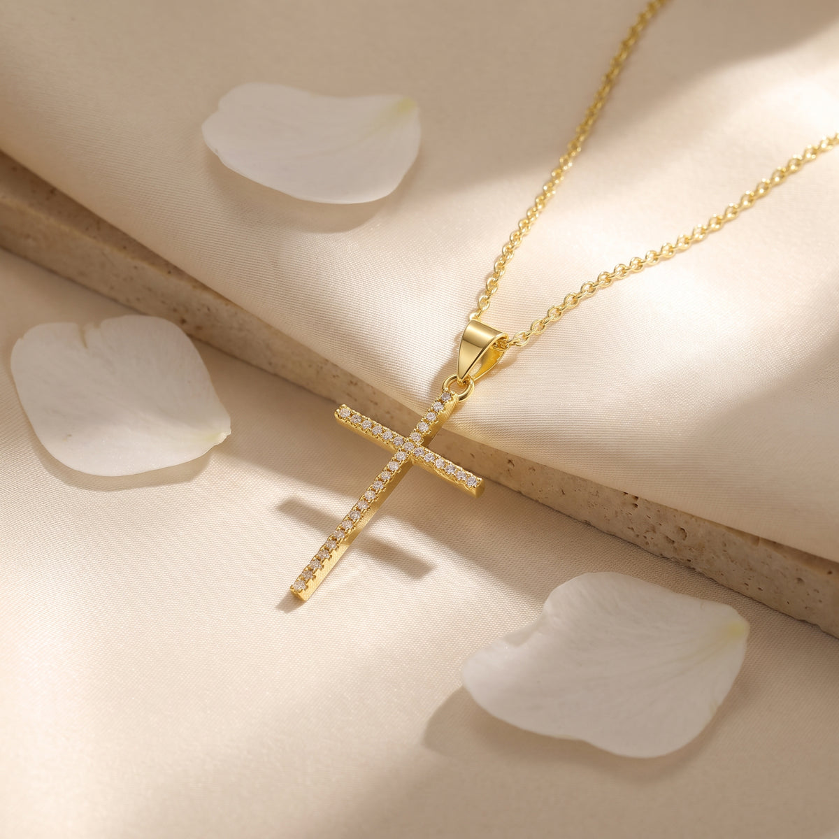 Gold-tone pavé cross pendant necklace resting flat on cream silk fabric alongside loose white petals.