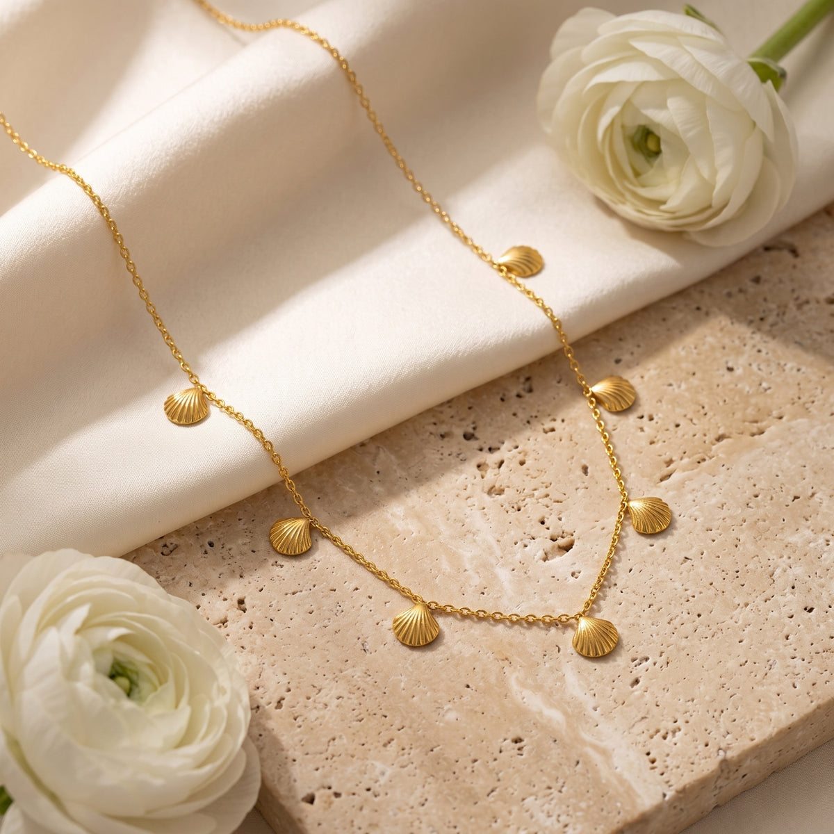Gold-tone scallop shell station necklace arranged across a textured beige slab beside white flowers.