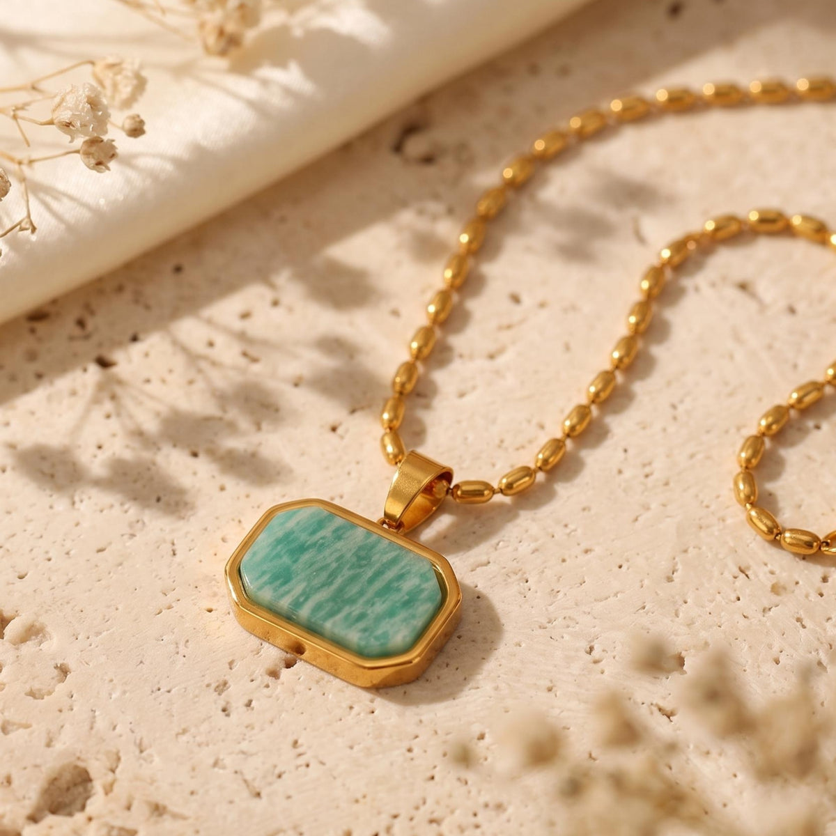Angled close-up of an octagonal green Amazonite pendant in a gold-tone frame on a textured beige surface.