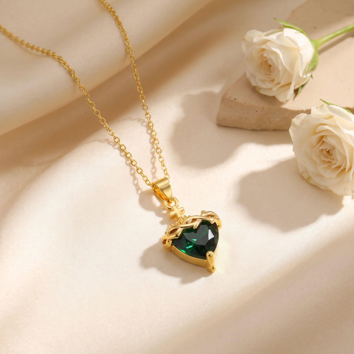 Angled view of a gold-tone necklace with a heart-cut simulated emerald pendant on white fabric beside white roses.