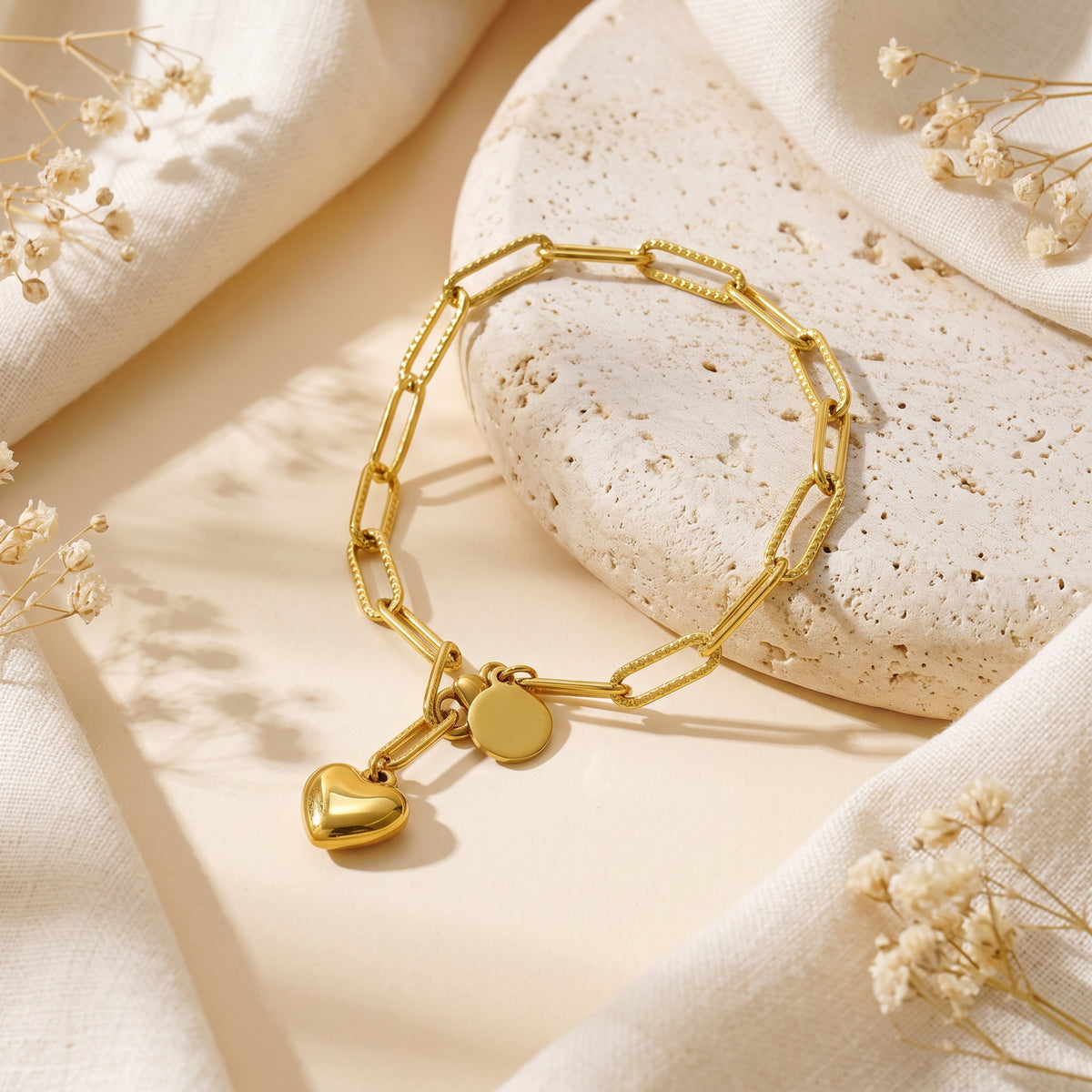 Angled view of the gold-tone heart charm link bracelet resting on travertine stone among fabric folds.