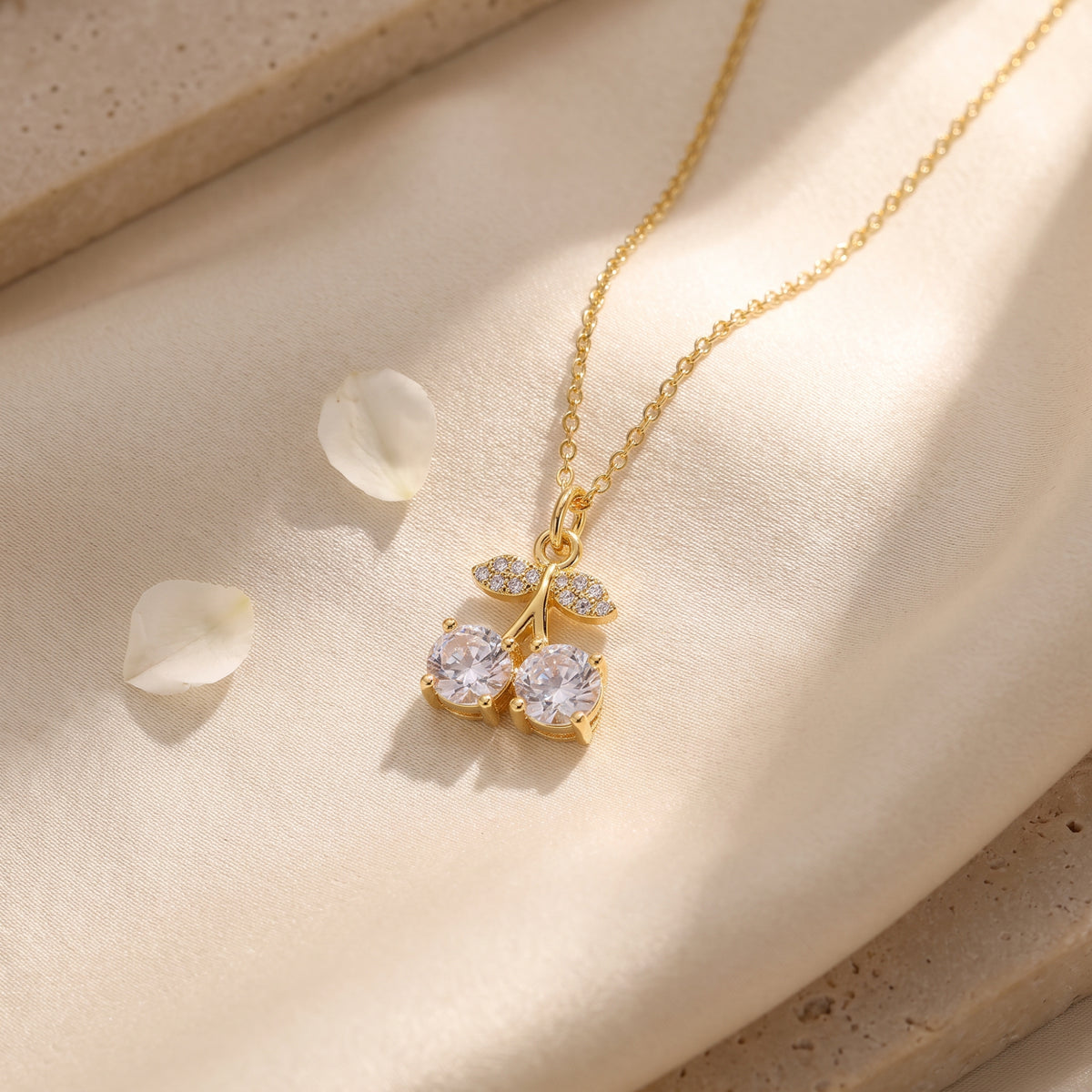 Flat lay of the gold-tone crystal cherry pendant necklace on folded cream fabric with flower petals.
