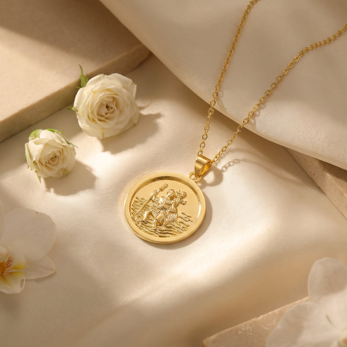 Gold-tone circular St. Christopher medallion necklace resting on beige fabric beside white roses.