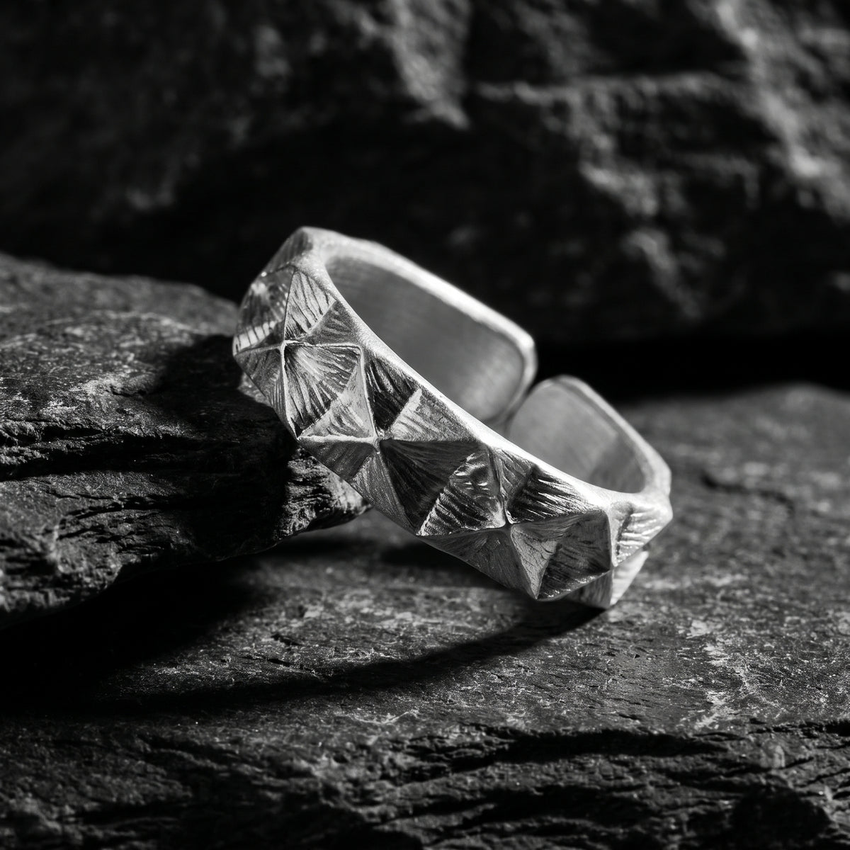 Angled silver faceted pyramid open-band ring resting on dark rock, with its opening visible behind the textured band.