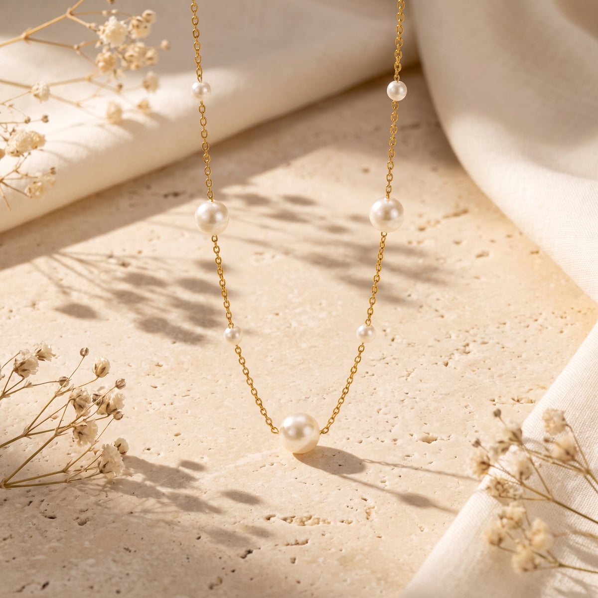 Gold-tone pearl station necklace arranged vertically on a textured stone surface with dried flowers.