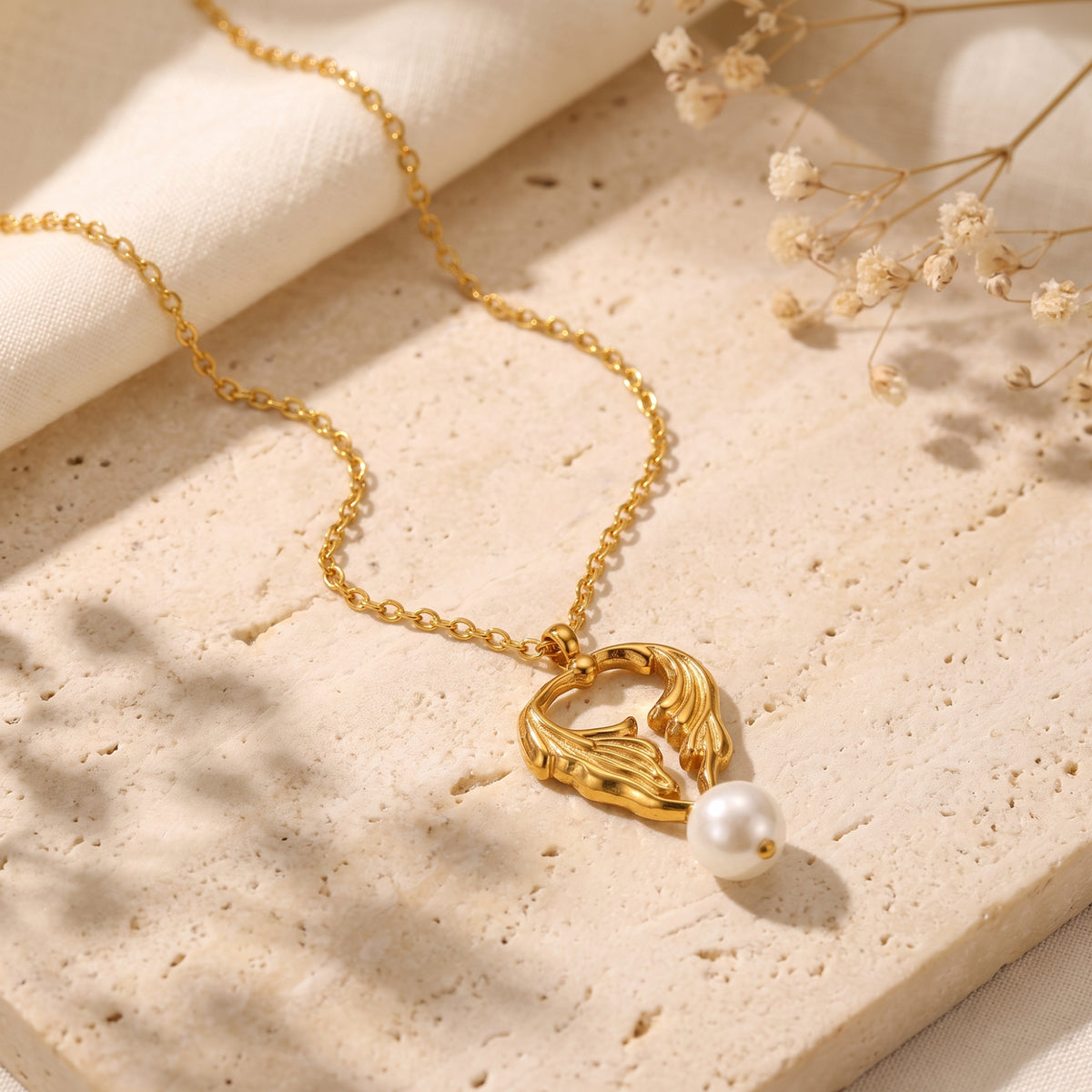 Gold-tone chain and leaf pendant with a white pearl drop arranged on a stone surface with dried flowers.