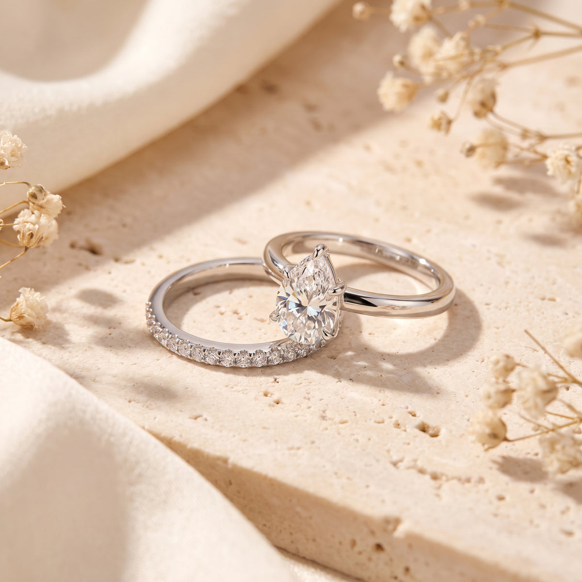 Two silver-toned rings overlap on a textured stone surface, showing a pear-cut solitaire and pavé band.