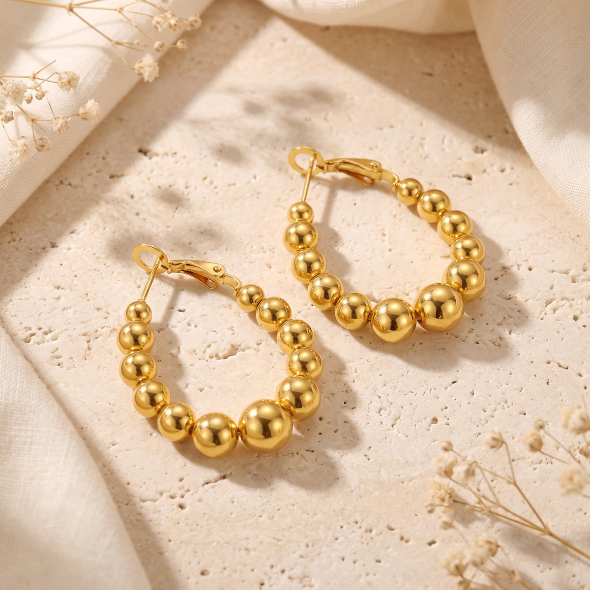 Two gold-colored graduated beaded hoop earrings lying flat on a textured stone surface.