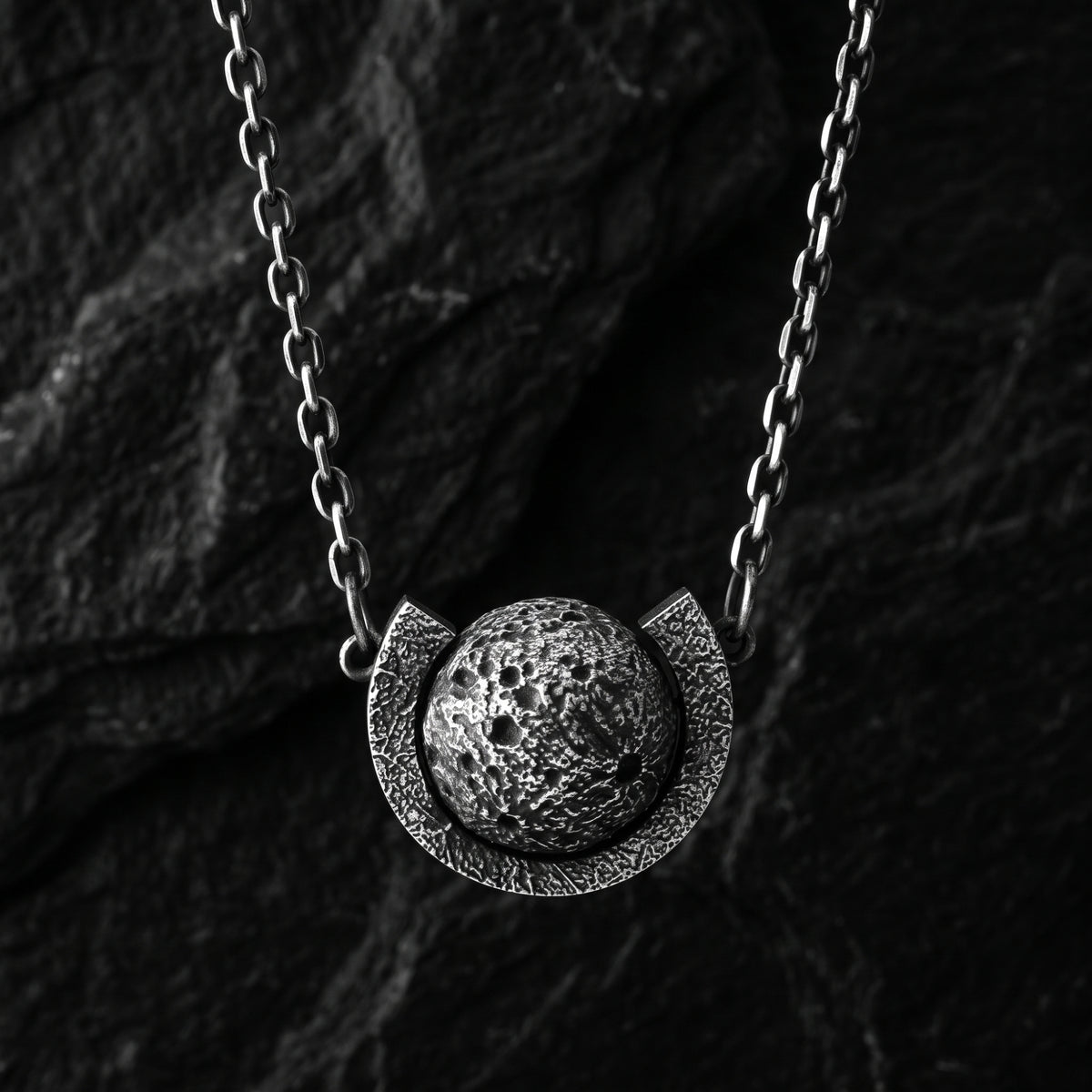 Frontal view of the silver cratered moon sphere pendant suspended within a textured arc on a dark background.