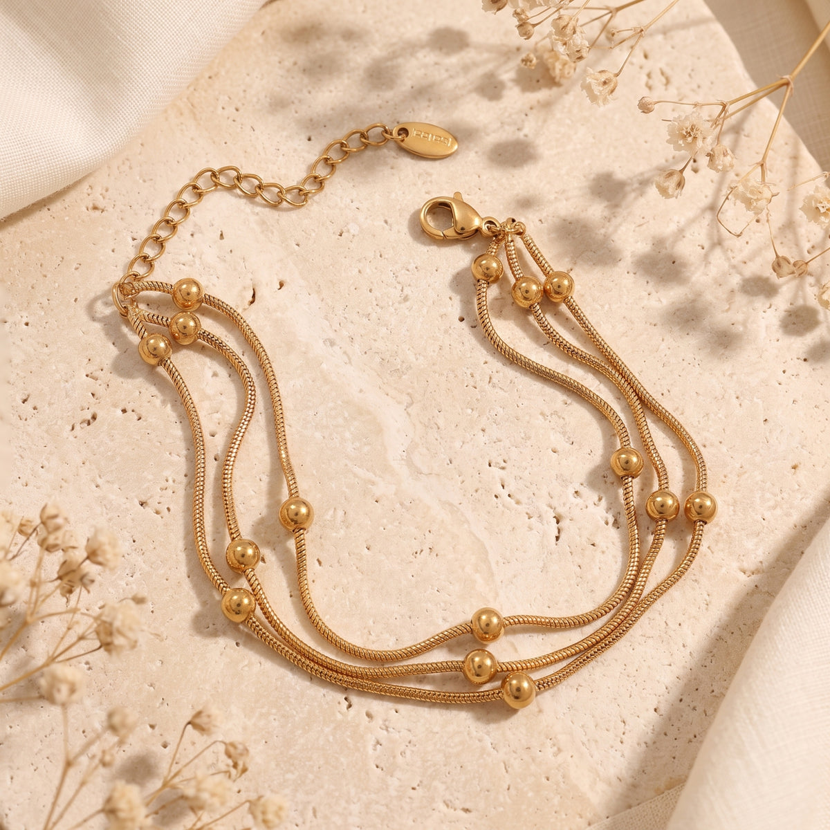Gold three-strand bead bracelet laid on beige stone, showing the clasp, extender chain, and logo tag.