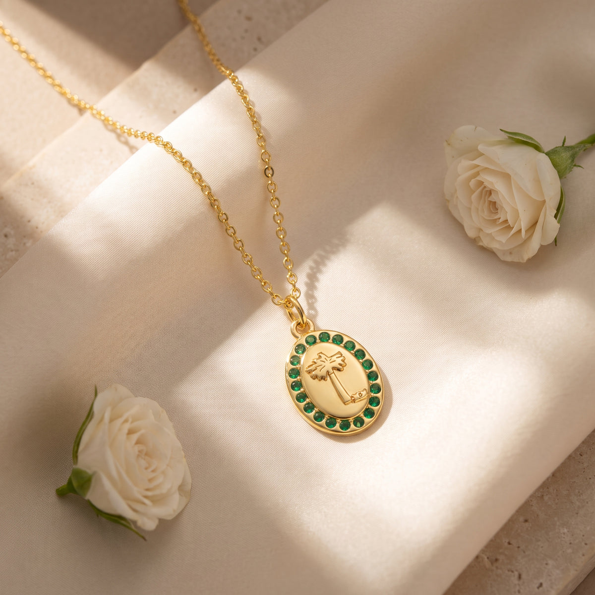 Gold-tone palm medallion necklace with green crystals on white satin beside two white roses.