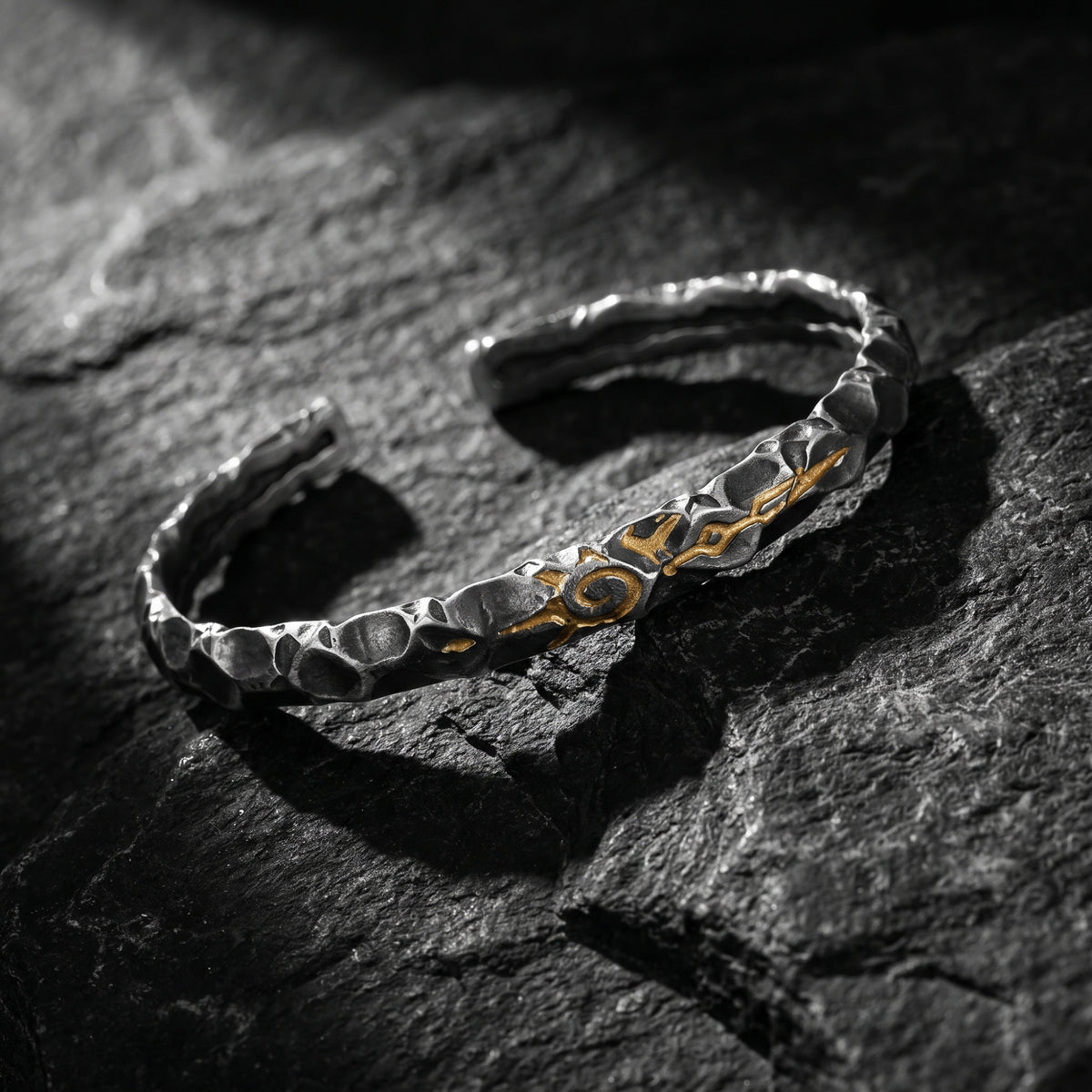 Angled view of the open hammered silver cuff bracelet with a gold-tone sigil on dark slate.