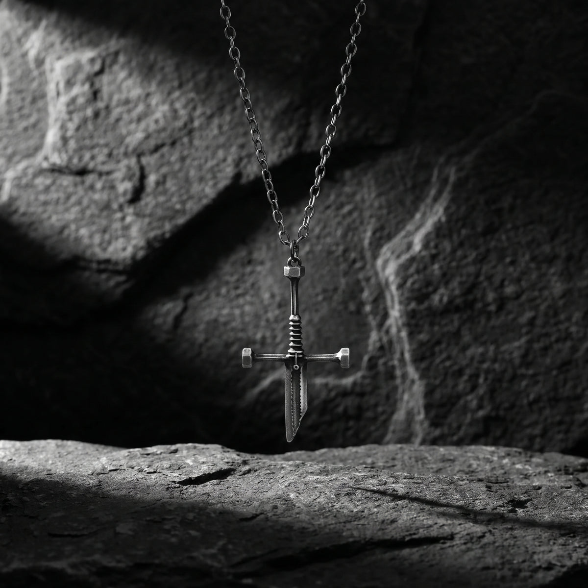 Silver dagger pendant and chain hanging vertically above a rough dark rock ledge.