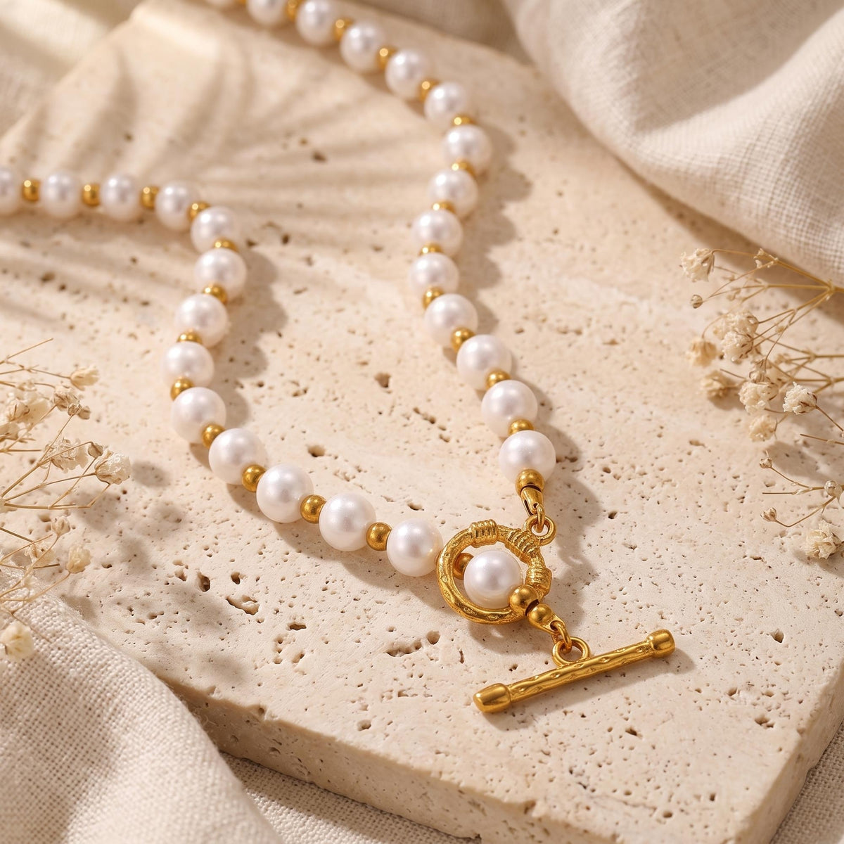 Flat-lay view of a white simulated pearl necklace with gold-tone spacer beads and a T-bar toggle clasp on a stone slab.