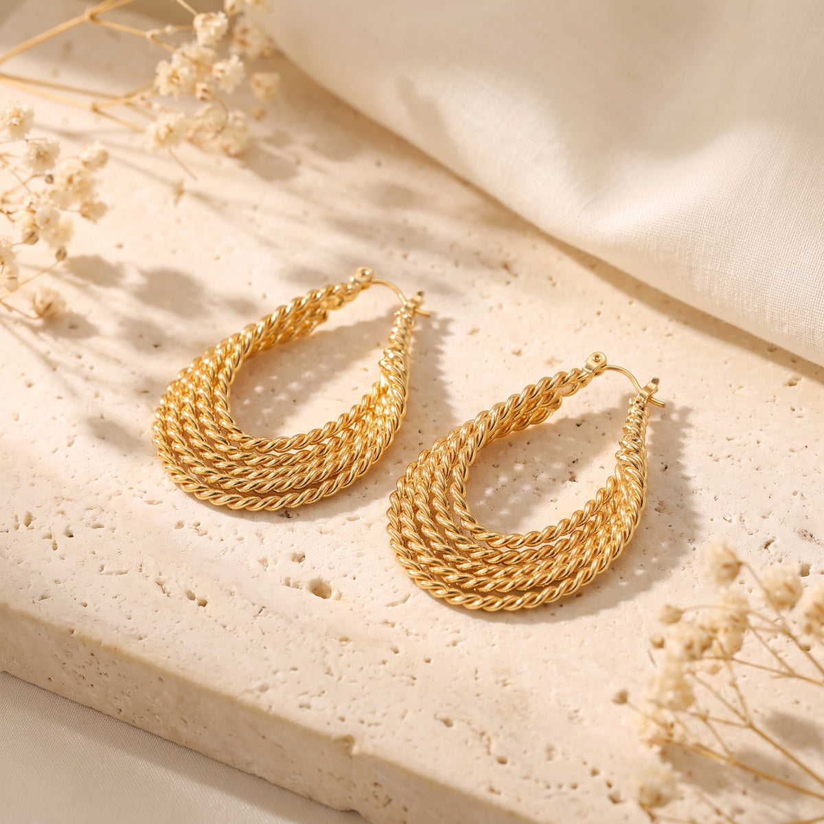 Angled overhead view of matching gold-tone twisted rope drop earrings lying on a cream stone slab.