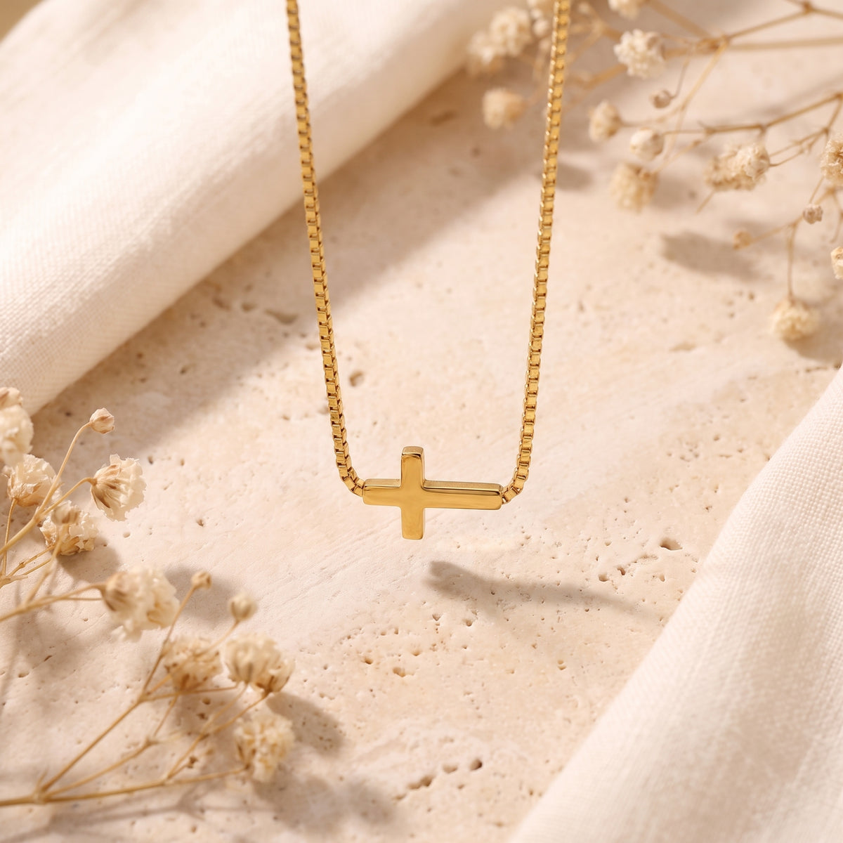Gold-tone sideways cross necklace laid on textured stone with dried flowers and white fabric.