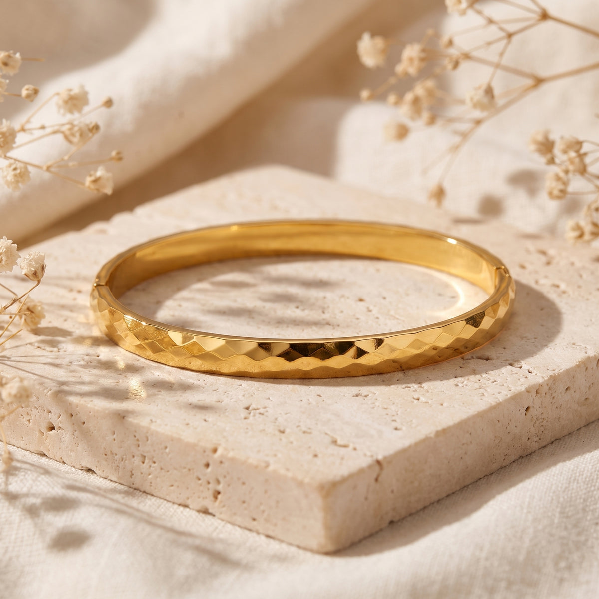Gold-toned faceted bangle bracelet lying flat on a square stone slab surrounded by dried flowers.