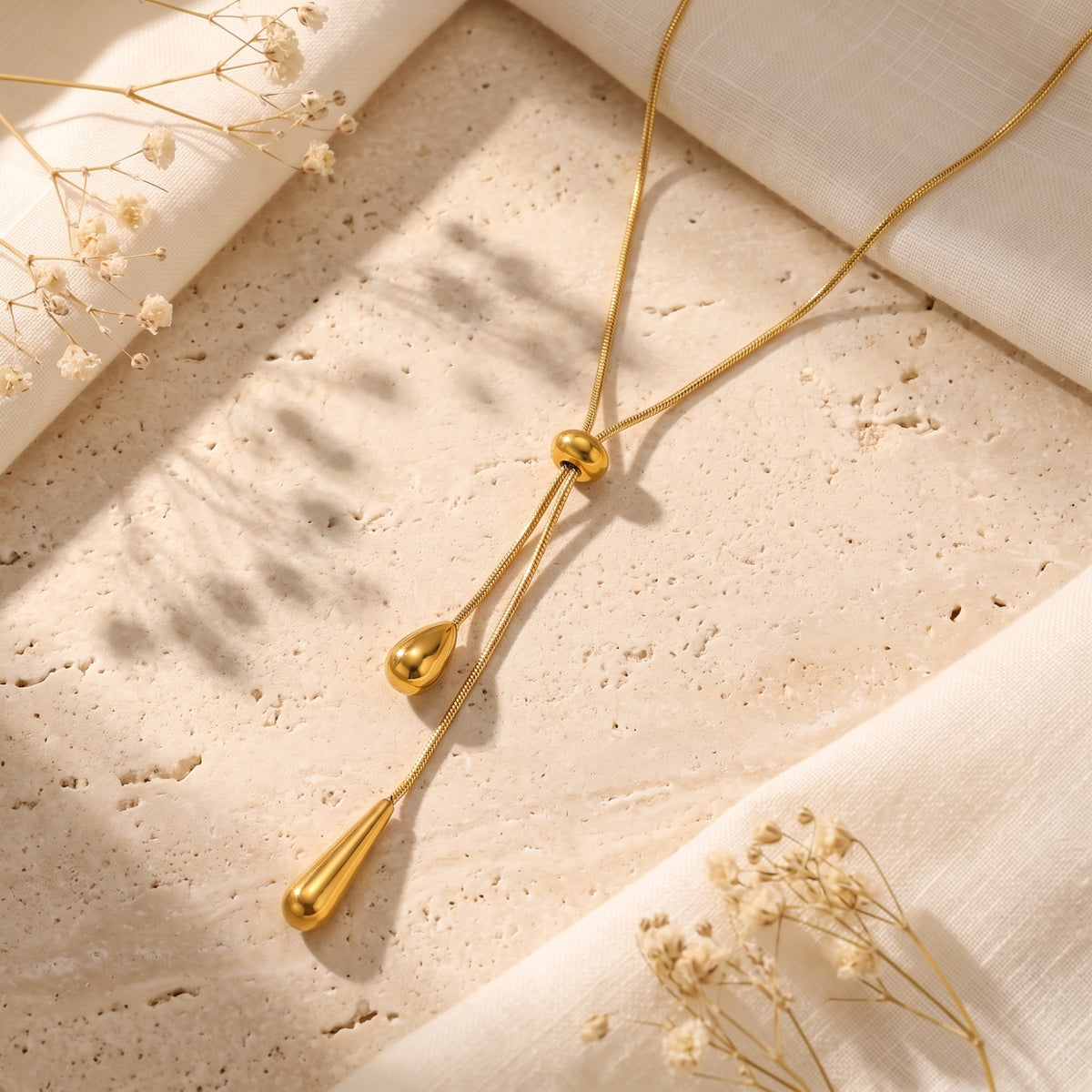 Flat lay of a gold-tone lariat necklace arranged diagonally on a travertine surface beside cream fabric and dried flowers.
