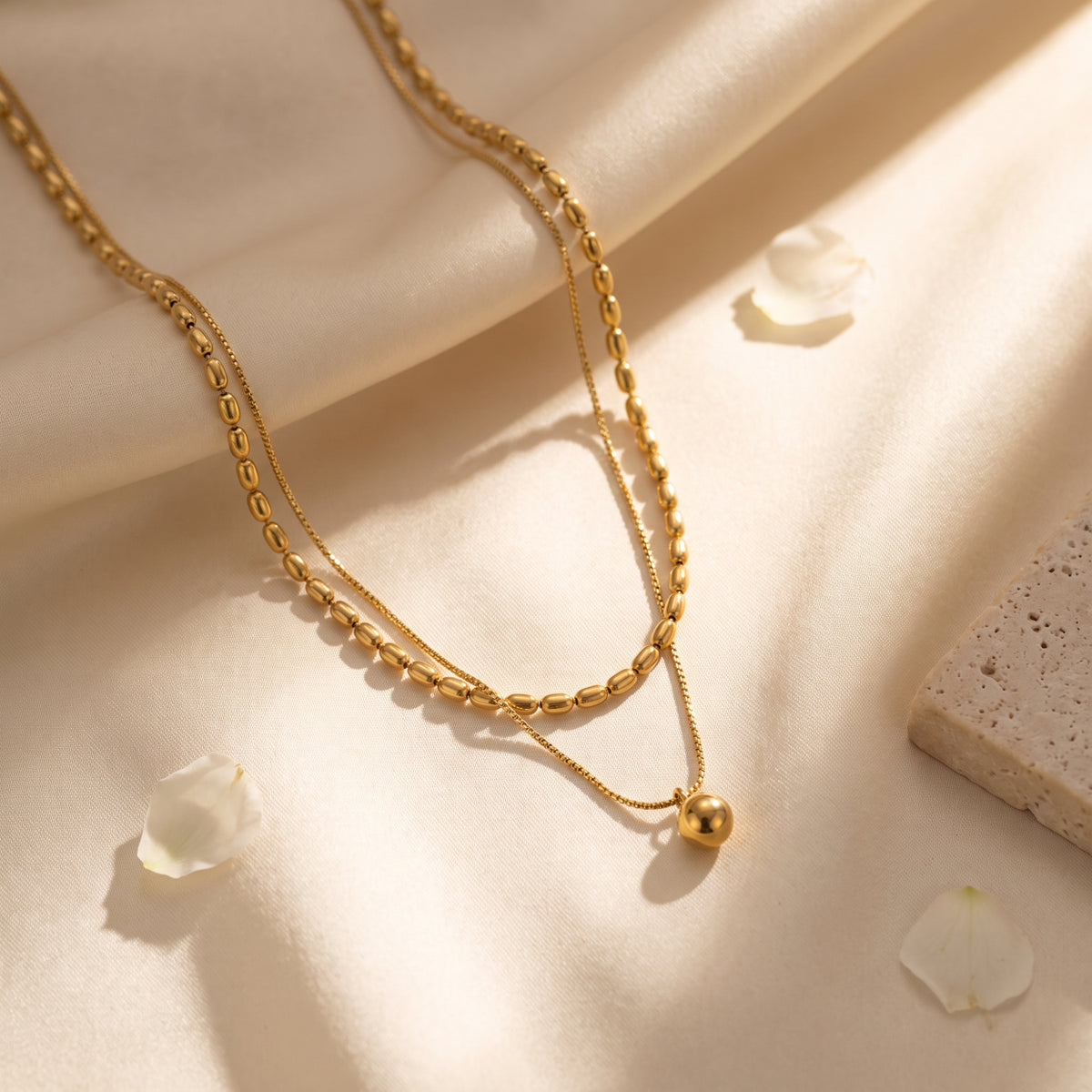 Gold-toned double-strand necklace lying on draped satin with delicate white flower petals.