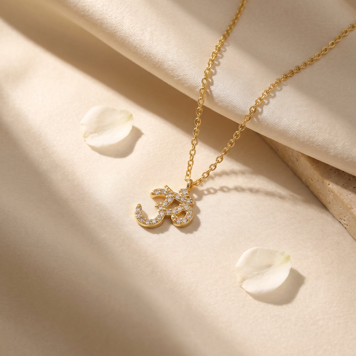 Gold-tone Om pendant necklace with clear stone accents resting on draped light fabric with flower petals.