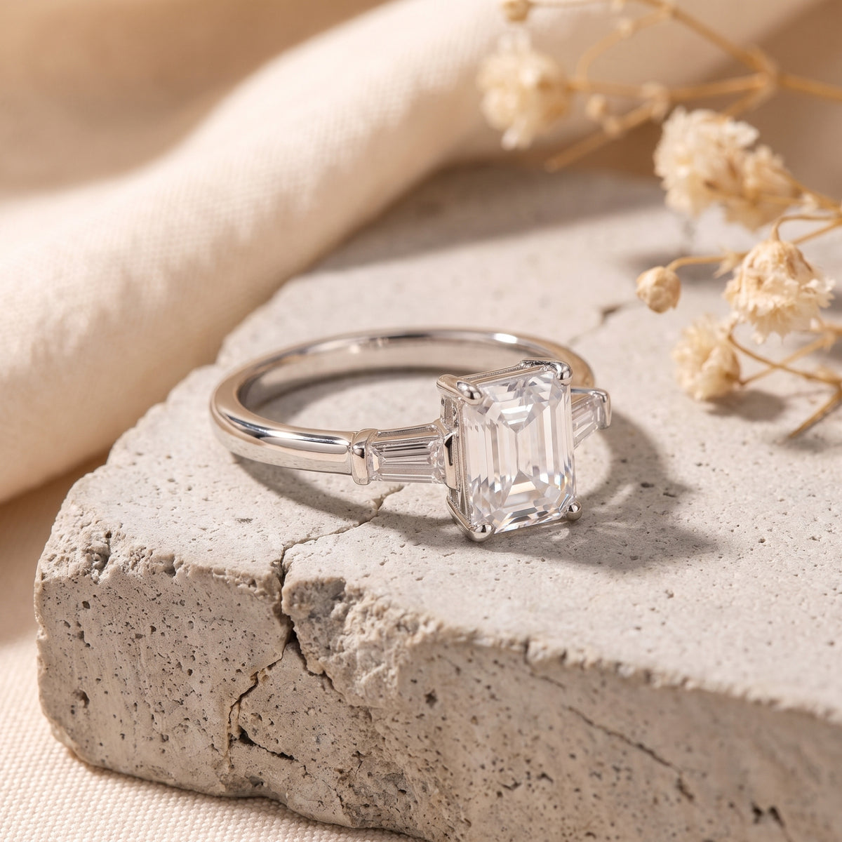 Angled view of a silver three-stone ring with an emerald-cut center stone resting on a gray stone slab.