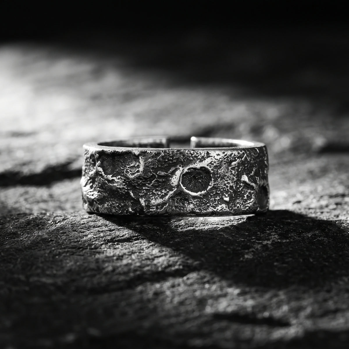 Front-facing close-up of a textured silver open-band ring displayed on a dark rock surface.