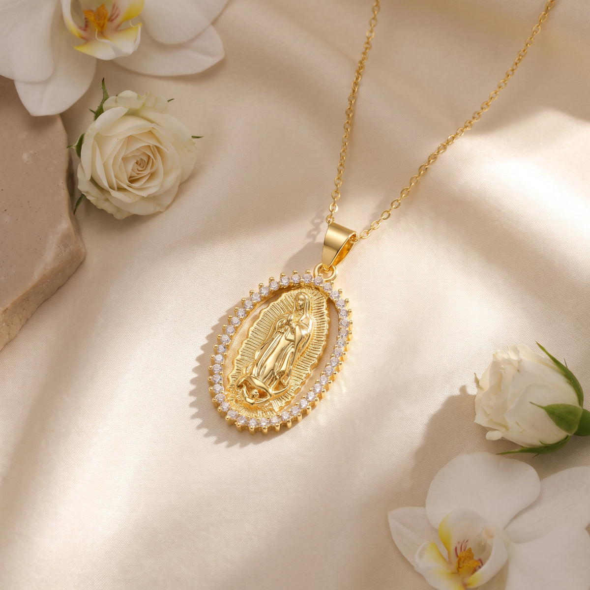 Flat lay of the gold-tone oval Guadalupe pendant necklace on beige satin, surrounded by white flowers.