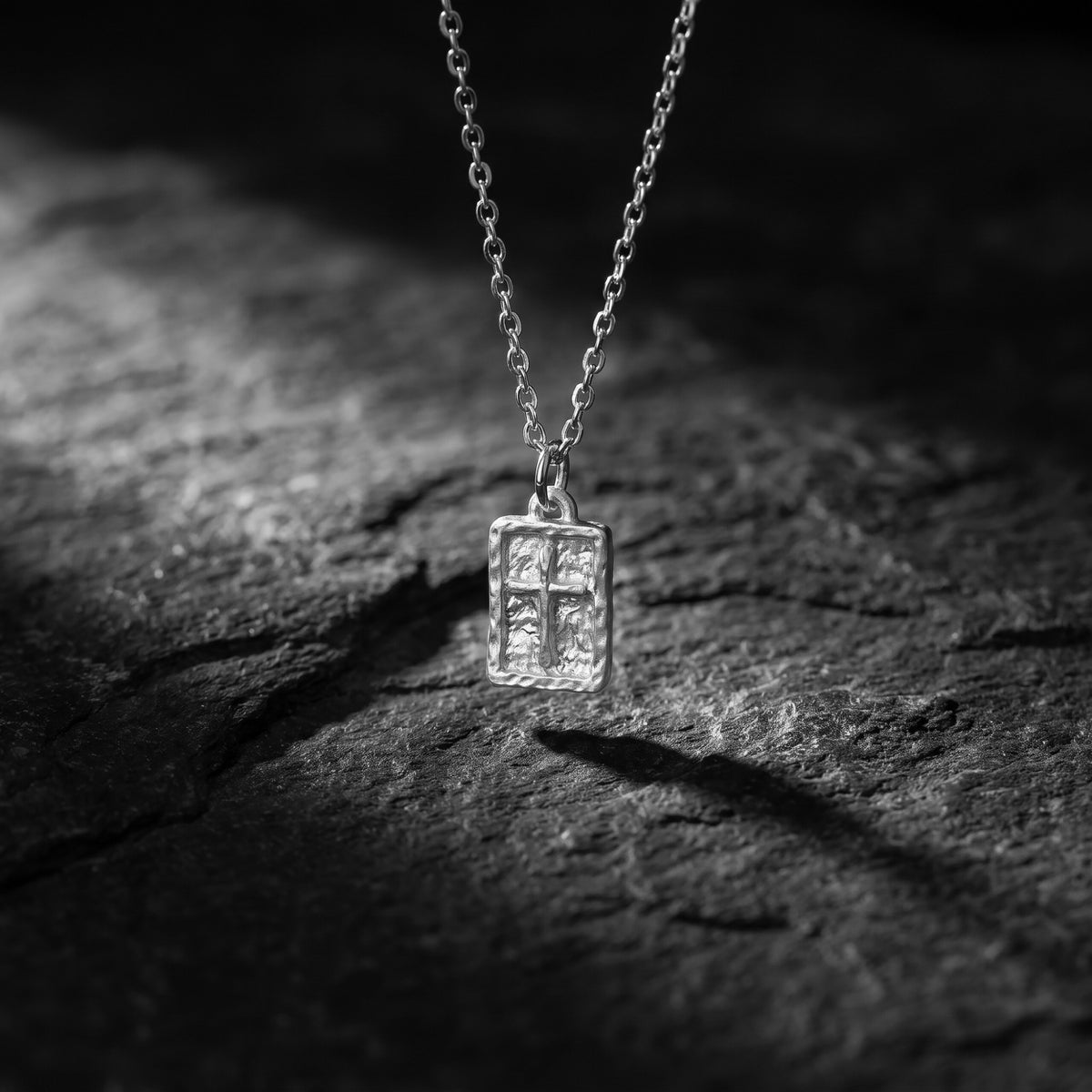 Silver textured cross pendant suspended vertically by a chain over a dark stone surface.