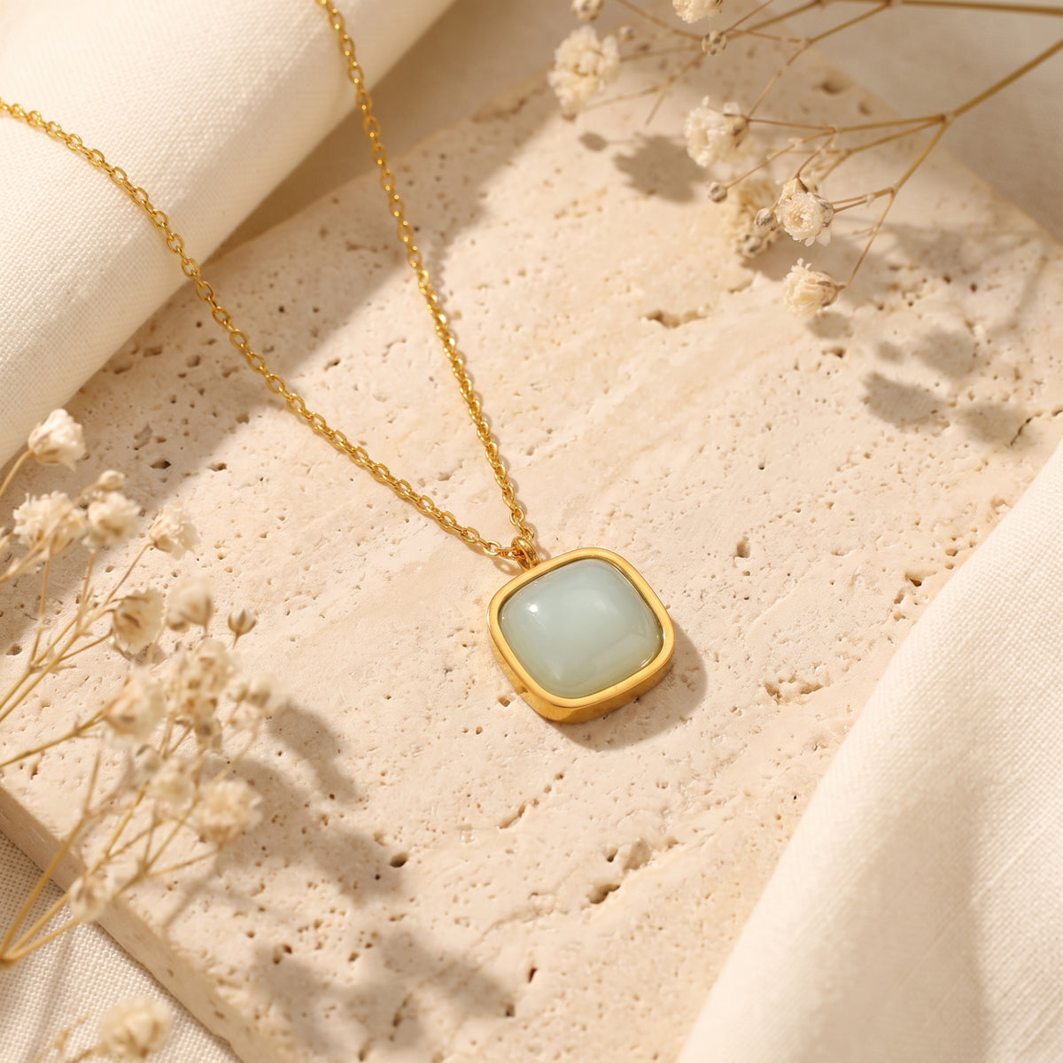 Angled overhead view of the gold-tone cushion bezel pendant necklace lying flat on a textured stone slab.