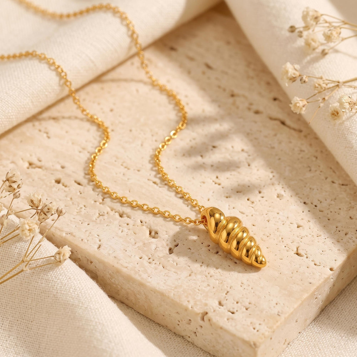 Gold-tone spiral shell pendant necklace arranged diagonally on a beige textured stone slab.