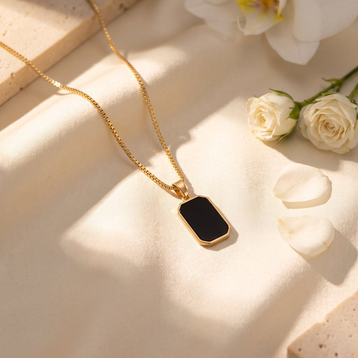 Gold-tone box chain necklace with an octagonal black pendant arranged diagonally on cream fabric beside white flowers.