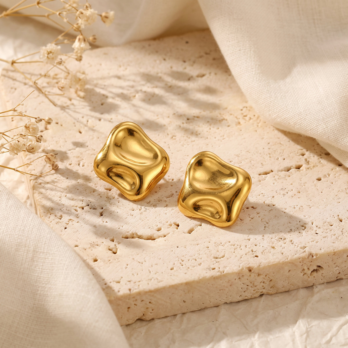 Angled view of two polished gold-tone sculptural square stud earrings on a textured beige stone slab.