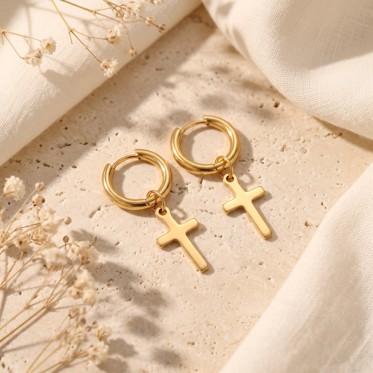 Pair of gold-tone cross drop hoop earrings lying flat on a beige stone surface beside soft fabric.