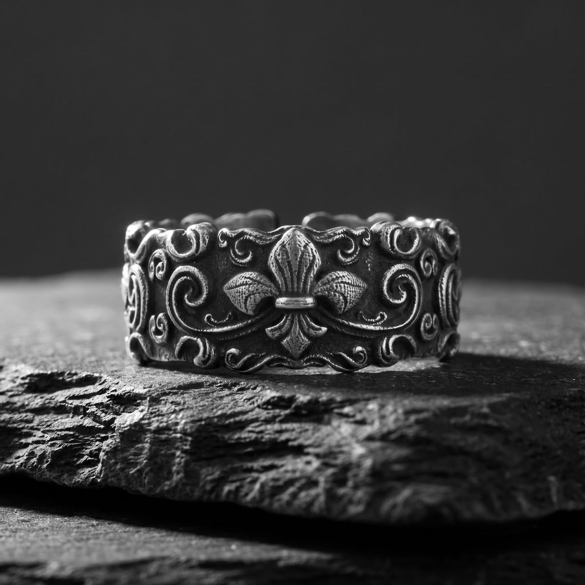 Front view of the silver ring resting on dark stone, showing its raised fleur-de-lis and scrollwork.