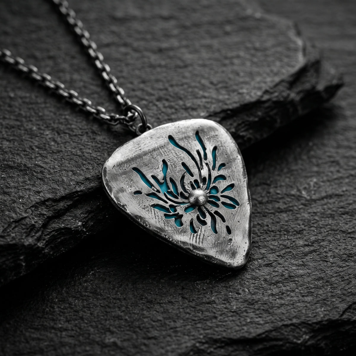 Angled close-up of the silver guitar-pick pendant on dark slate, showing its floral cutout and cyan backdrop.
