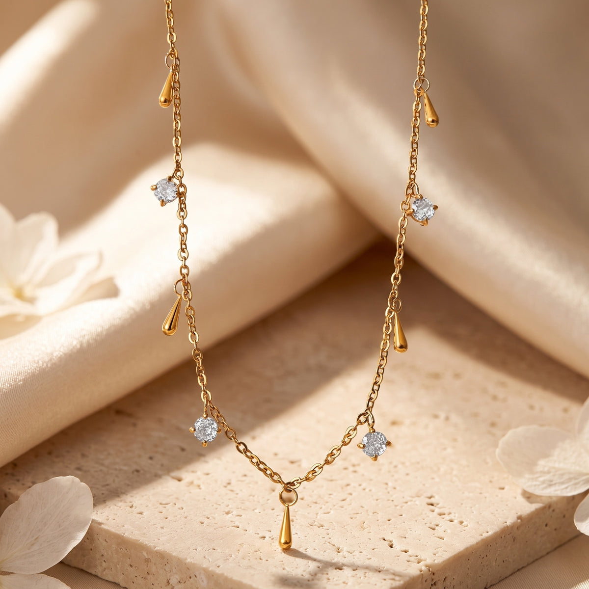 Gold-tone droplet station necklace arranged vertically on cream fabric and a stone slab, with clear round stones.