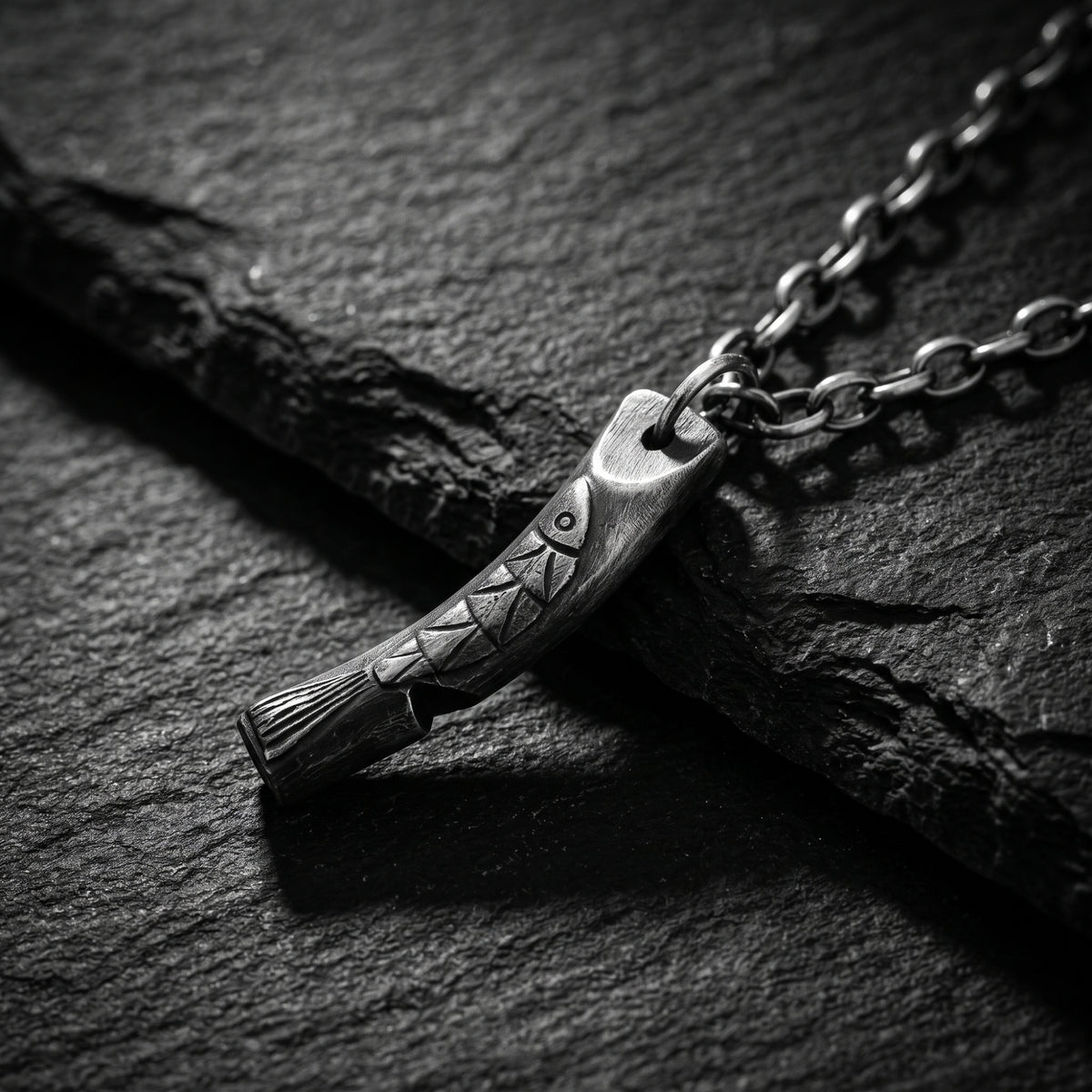 Angled silver carved fish bar pendant resting on a dark slate surface with its chain extending behind it.